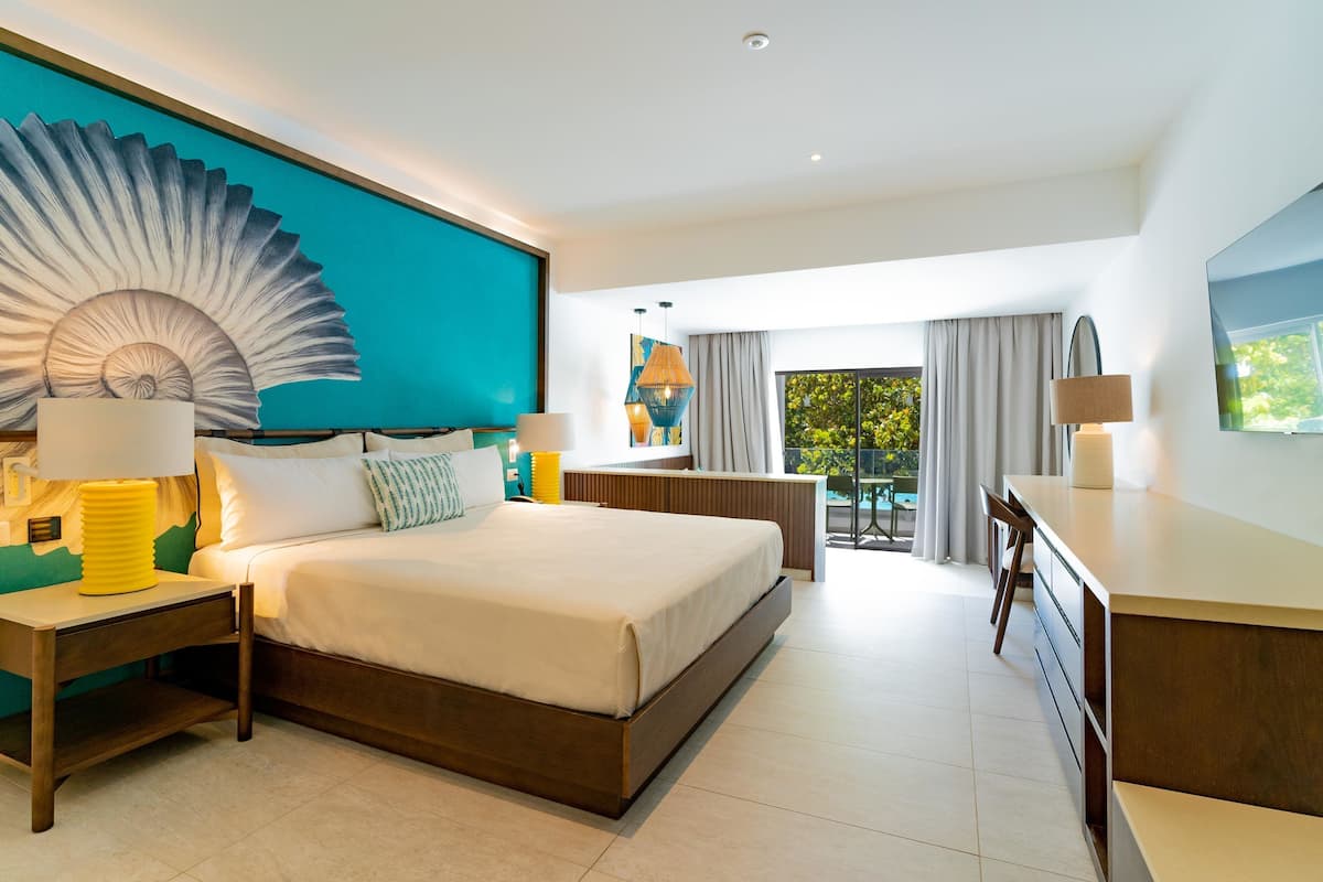 A modern hotel room with a large bed, a desk, and a balcony with a view of greenery.