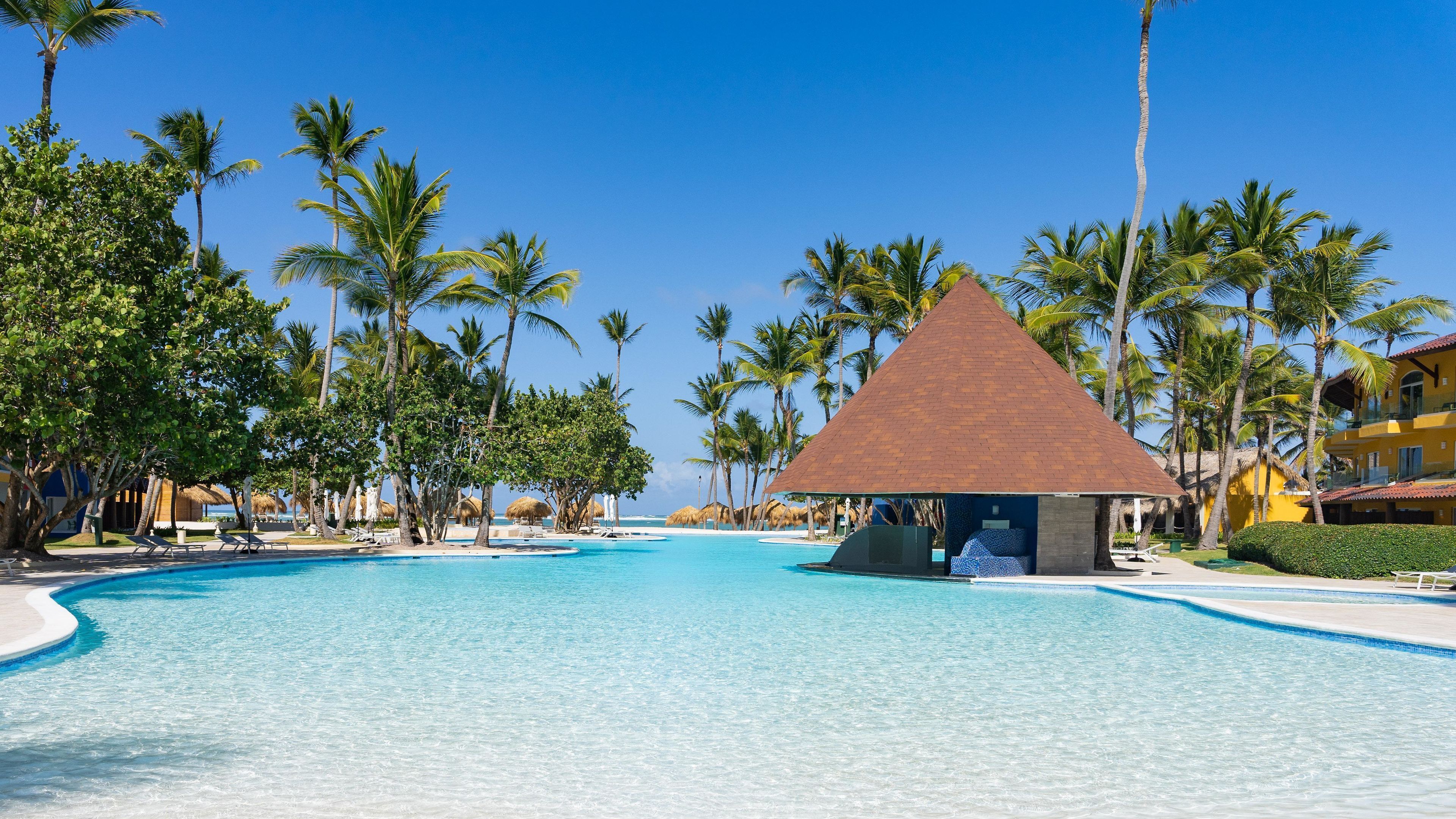 Une piscine d’un complexe hôtelier, avec une eau d’un bleu limpide, entourée de palmiers et bordée d’une structure à toit de chaume.
