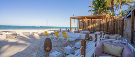 Un salon de plage doté d’une terrasse en bois, de chaises longues blanches et jaunes, et d’un mur en pierre en arrière-plan.