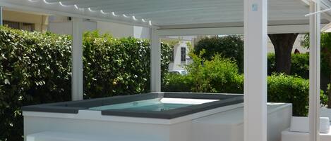 A covered outdoor swimming pool with steps leading into it, surrounded by greenery and a white building in the background.