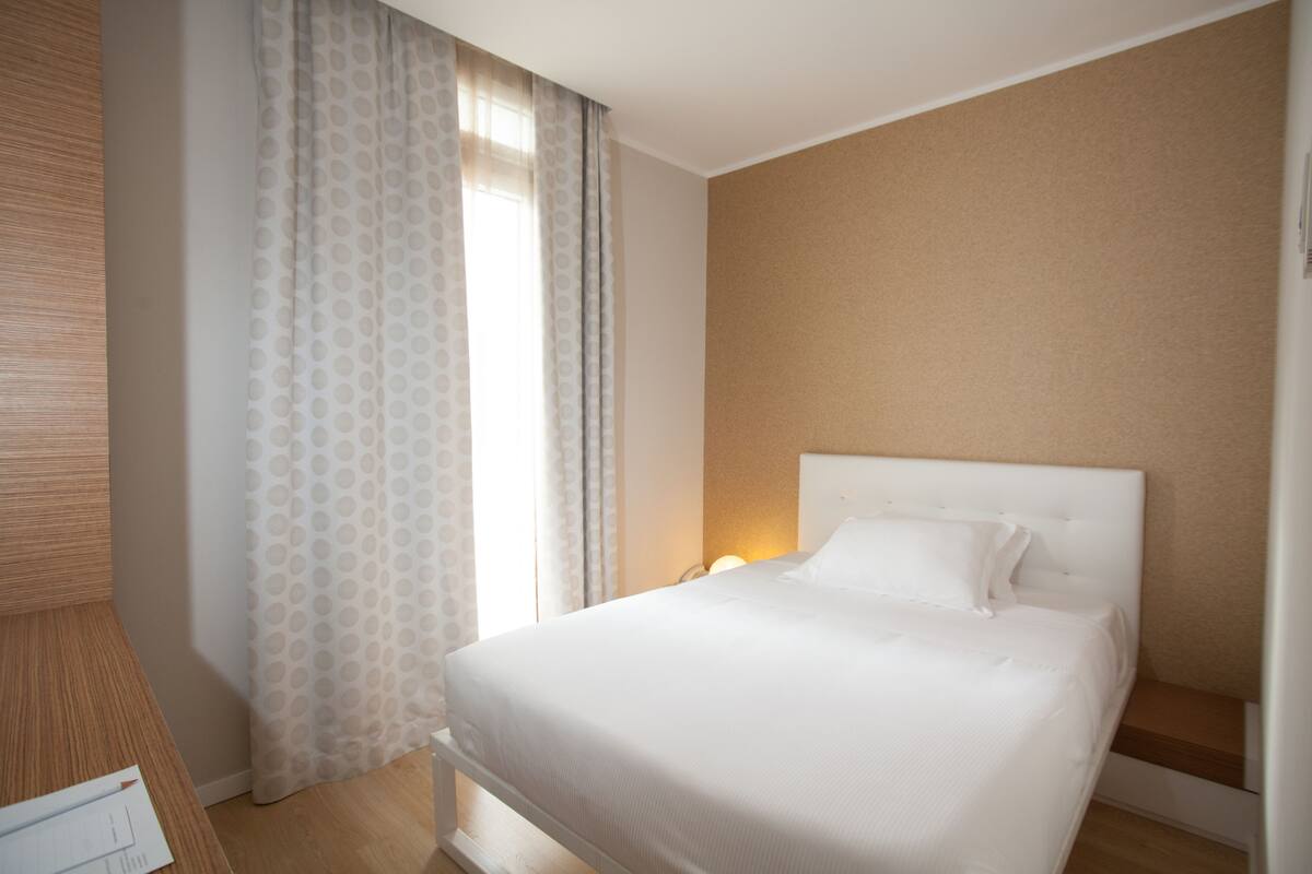 A hotel room with a large bed, a wooden nightstand, a desk, and a window with curtains.