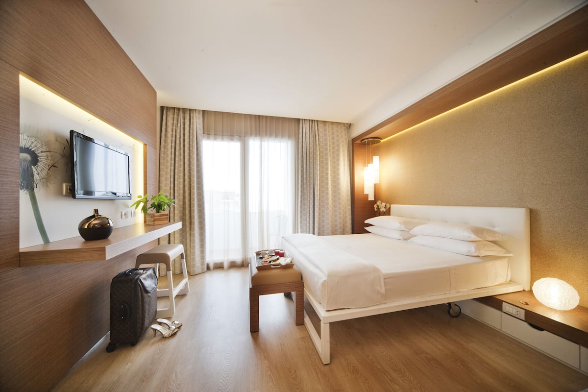 A modern hotel room with a large bed, a desk with a flat-screen TV, a bedside table with a lamp, and a window with curtains.