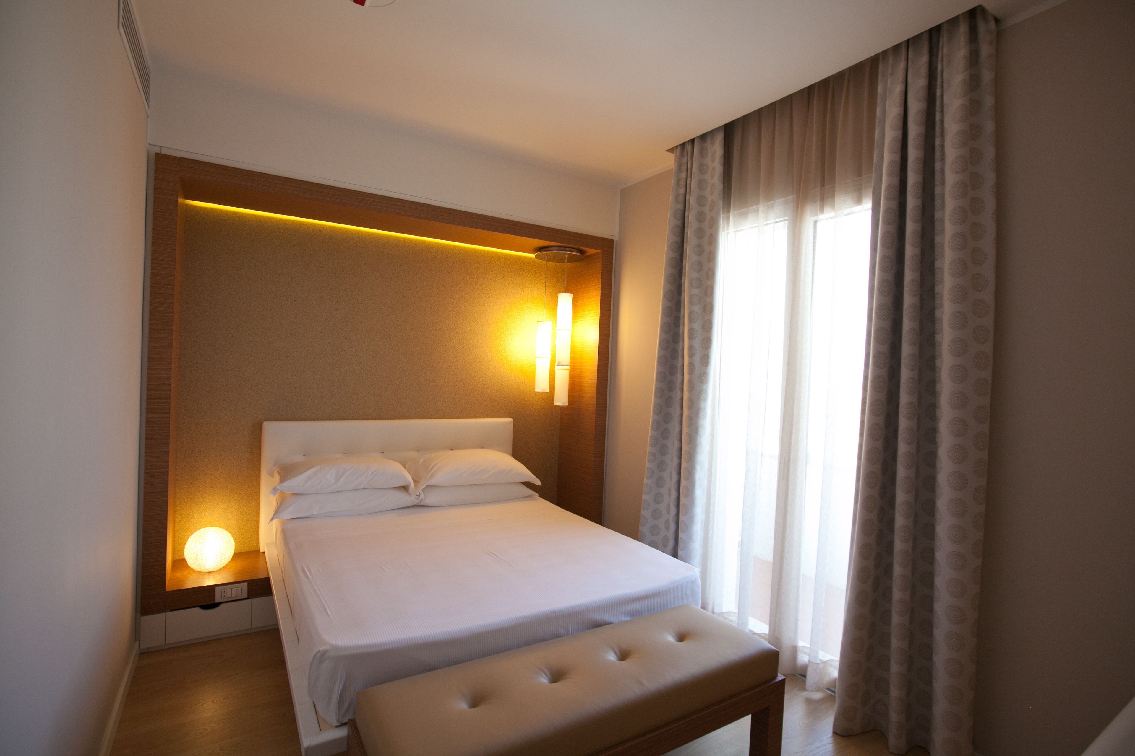 A hotel room with a bed, a bedside lamp, a window with curtains, and a wall-mounted light fixture.