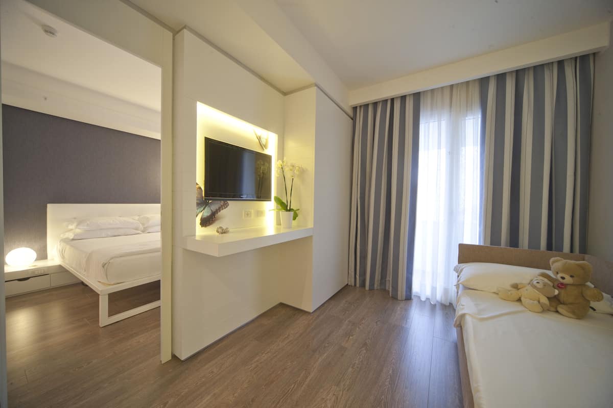 A hotel room with a bed, a bedside table with a TV, a small desk with a plant, and a window with curtains.