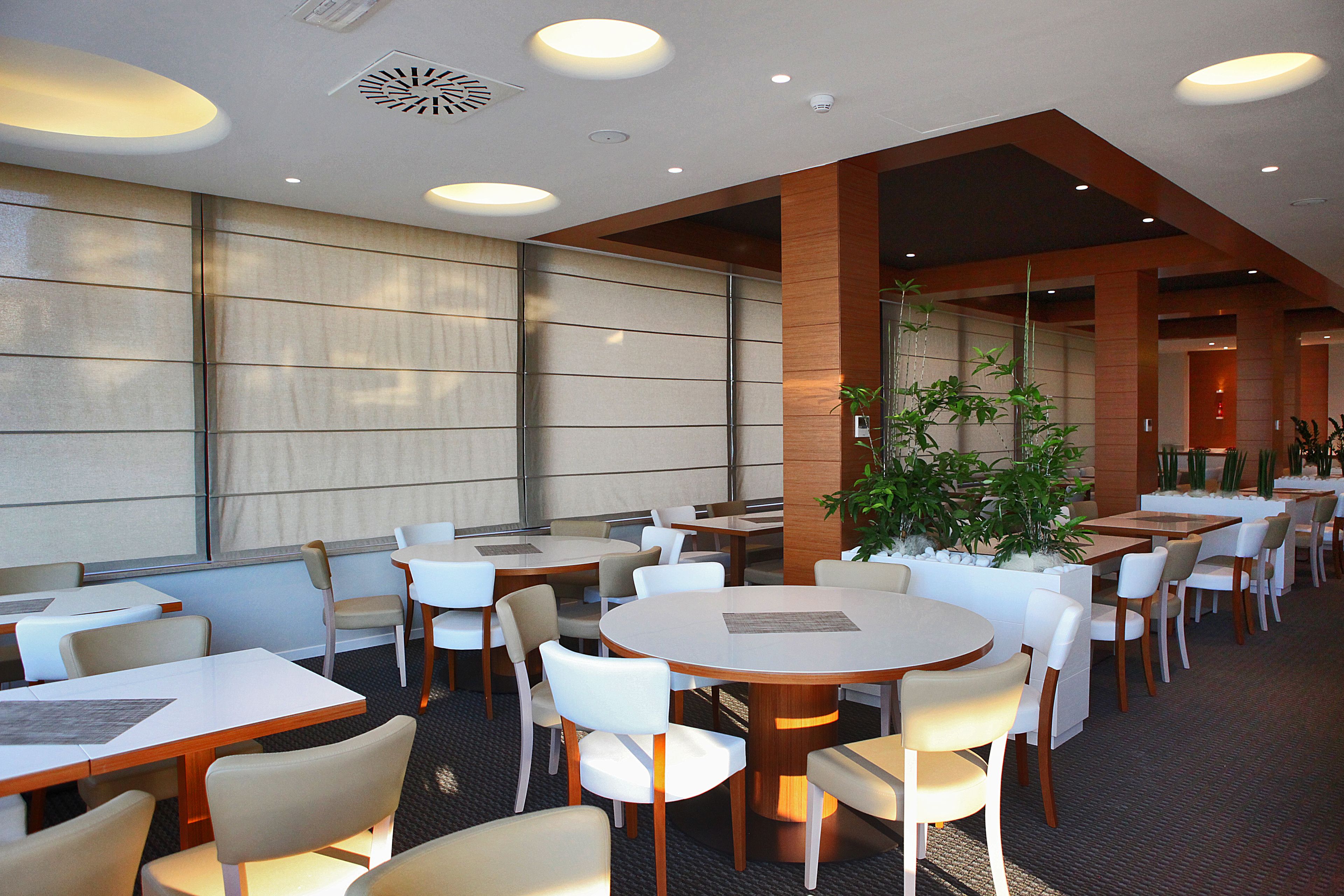 Un restaurante con mesas redondas y sillas blancas, ventanas amplias y una planta en maceta.