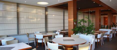 A restaurant with round tables and white chairs, large windows, and a potted plant.