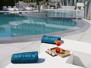 Zona junto a la piscina con sillones reclinables, una toalla con la marca 'OXYGEN' y una bandeja con bebidas y aperitivos.