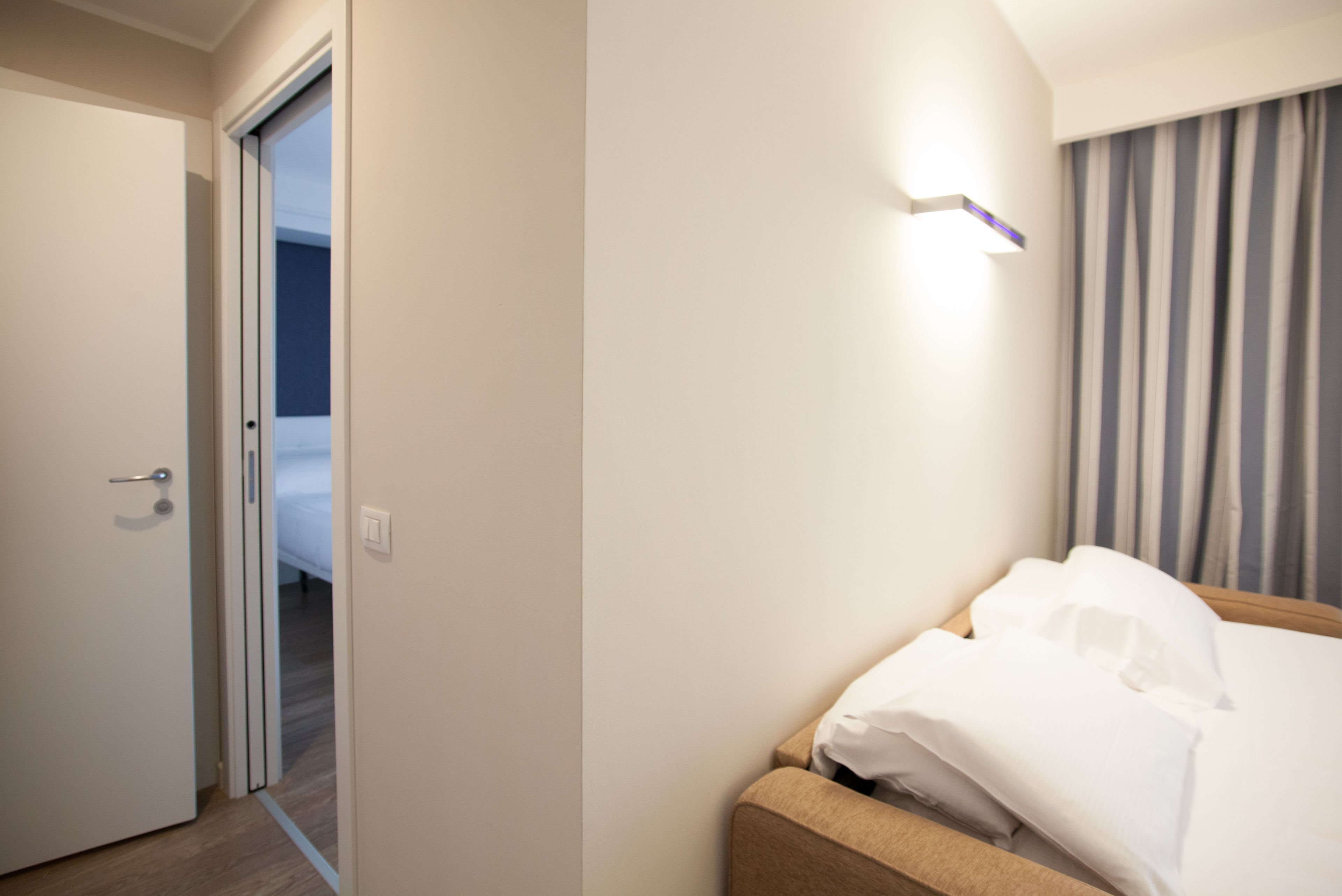 A hotel room with a bed, a door, and a light fixture on the wall.