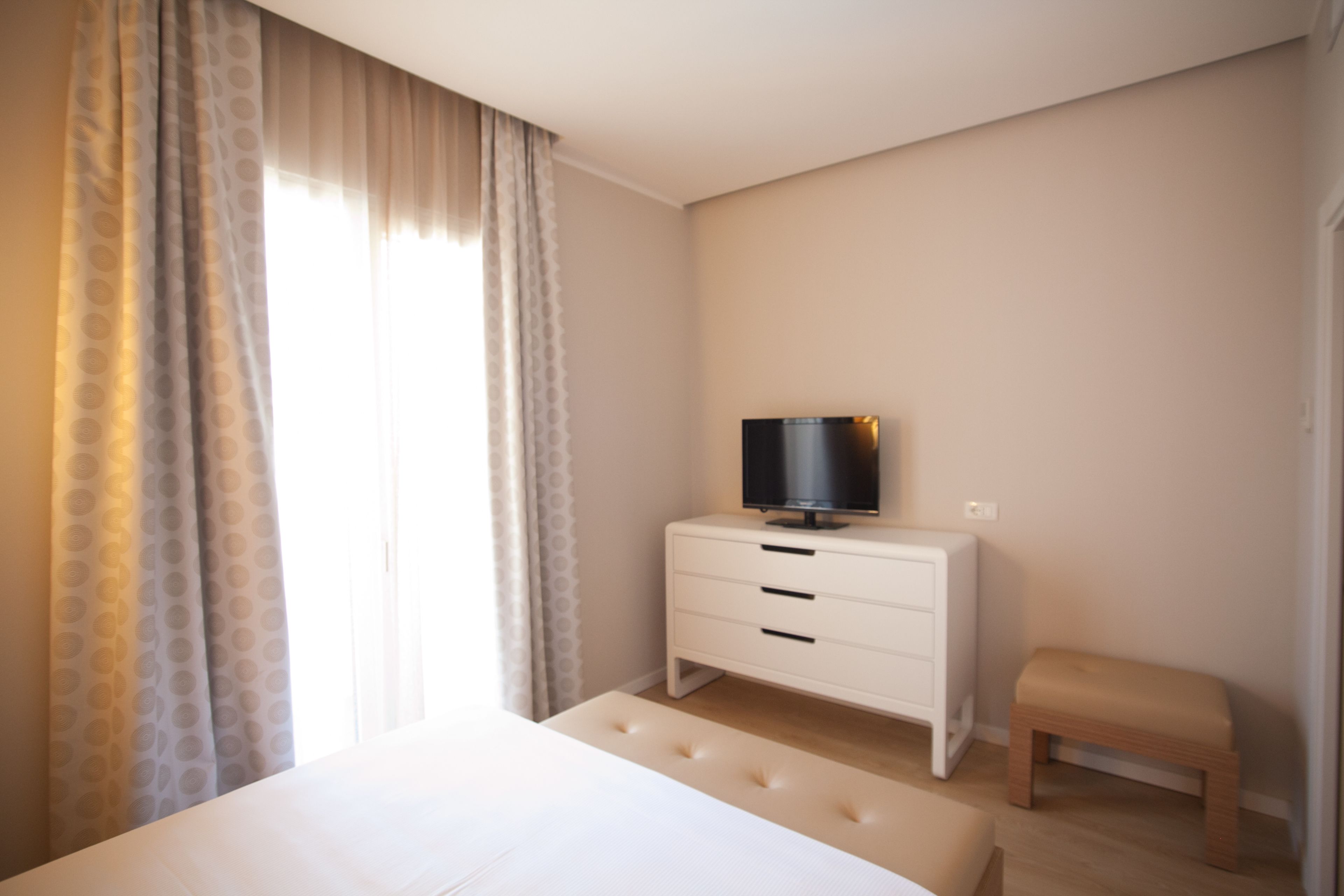 Un dormitorio con una cama, un televisor sobre un tocador blanco, un banco de madera y una ventana con cortinas.