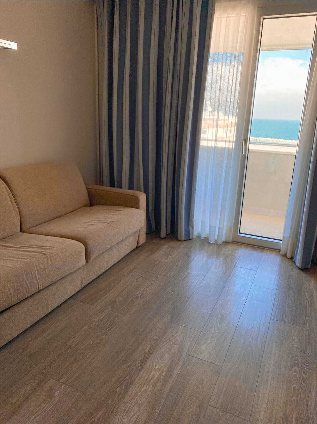 A living room with a beige sofa, wooden flooring, and a view of the ocean through a glass door.