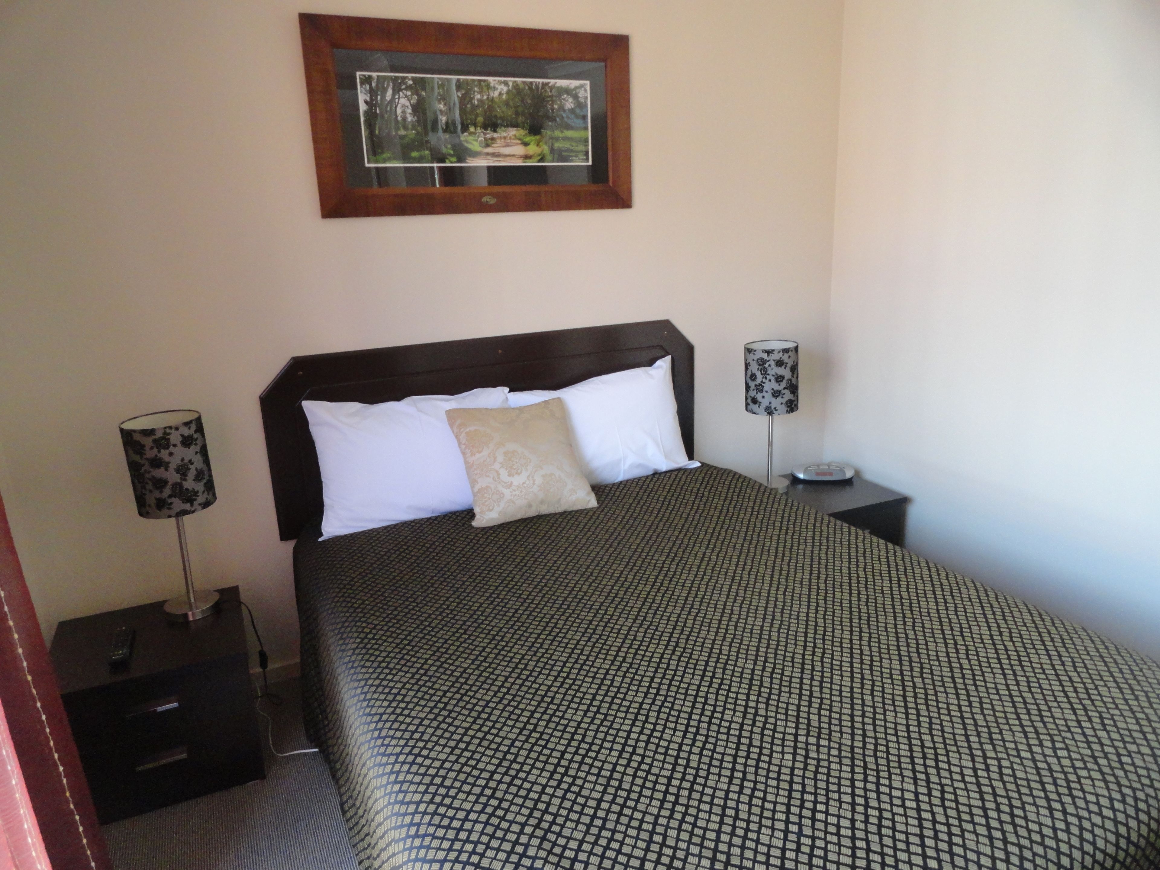 A neatly made bed with a patterned bedspread, two bedside lamps, a small bedside table, and a framed picture on the wall.