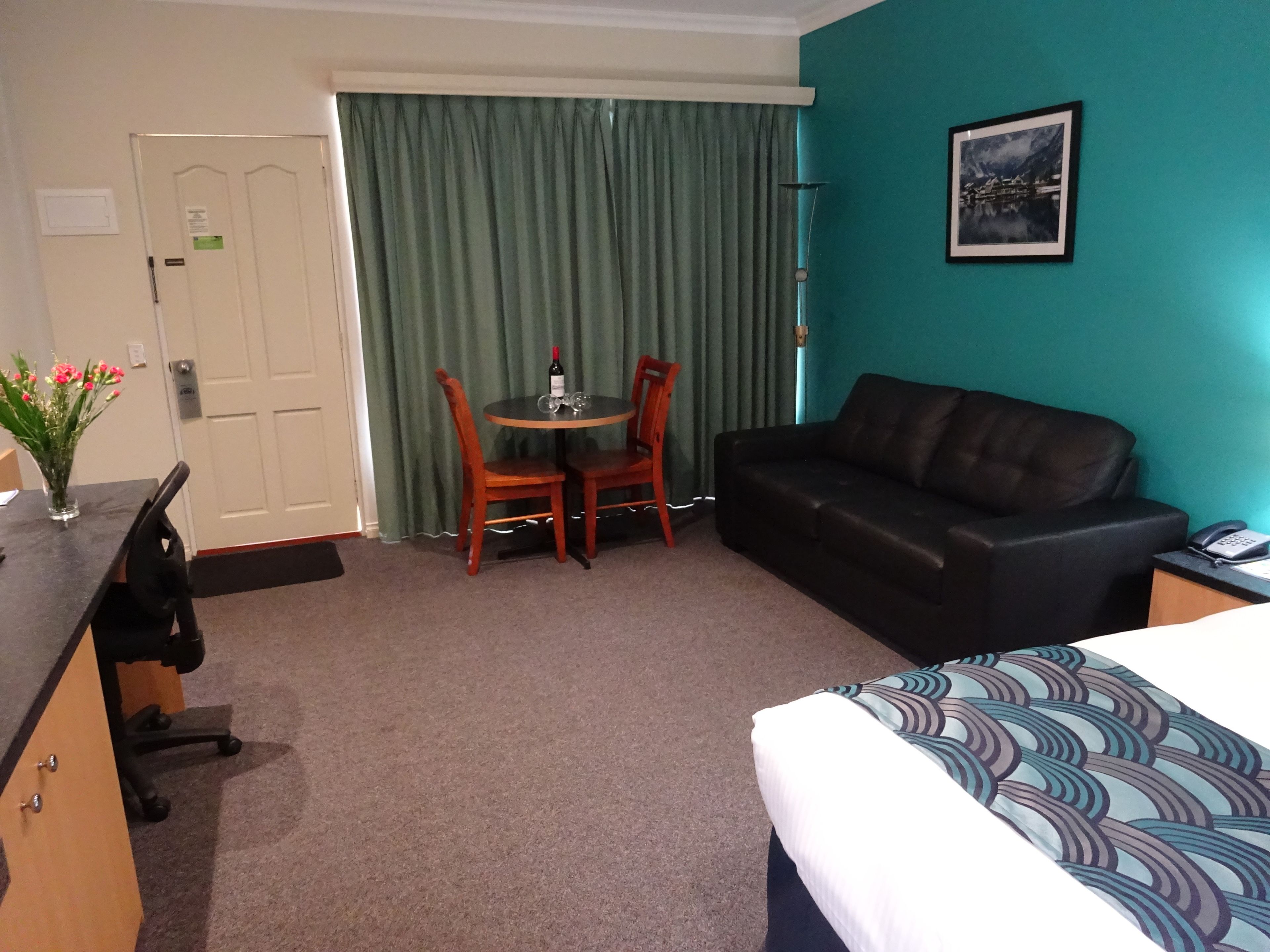 A hotel room with a bed, a desk, a chair, a table with a bottle of wine, a couch, and a painting on the wall.
