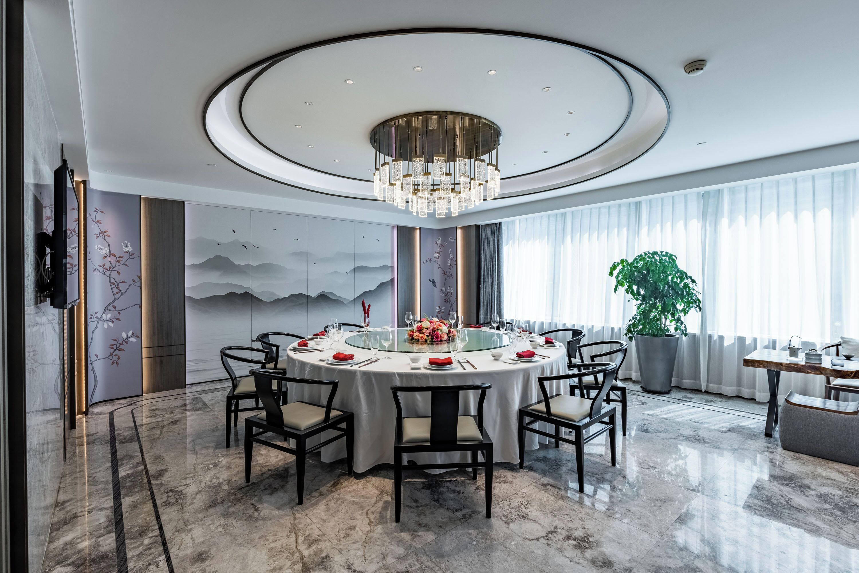 A modern dining room with a round table, chairs, a chandelier, and a large potted plant.