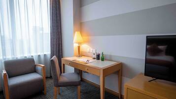 A hotel room with a desk, chair, television, lamp, and a window with curtains.
