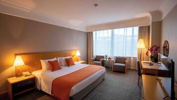 A hotel room with a large bed, two bedside tables with lamps, a desk with a computer, a chair, and a small table with a vase of flowers.