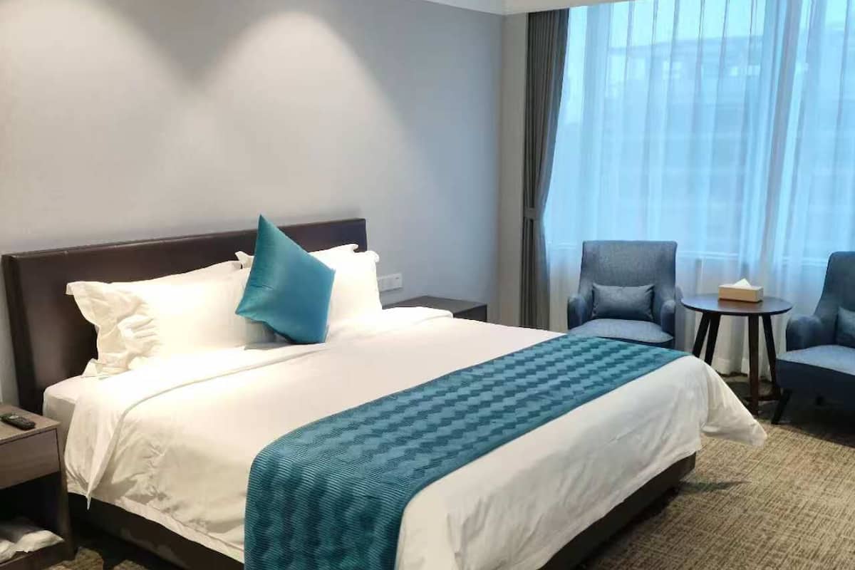 Een hotelkamer met een groot bed, een nachtkastje, een stoel, een klein tafeltje en een raam met gordijnen.