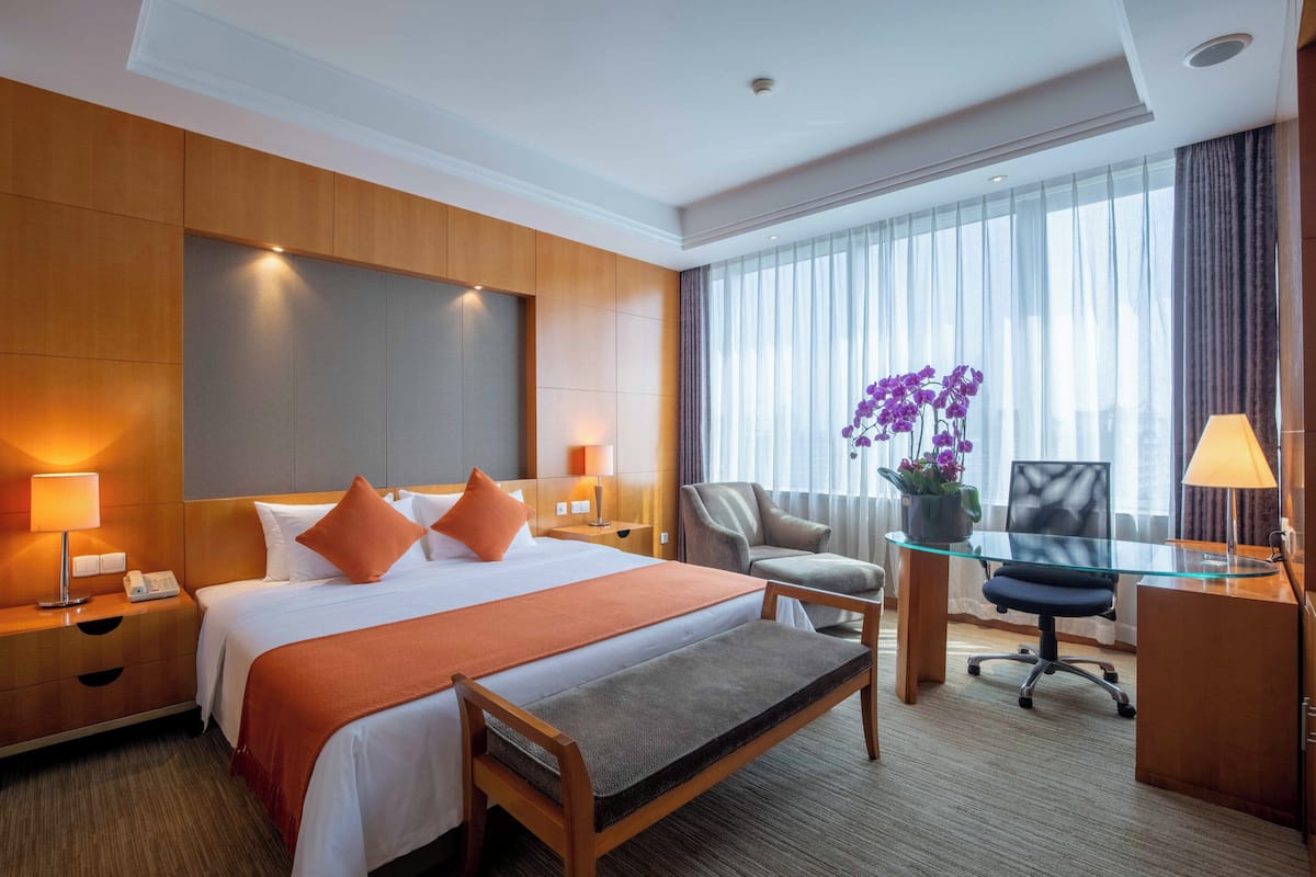 Een hotelkamer met een groot bed, een bureau, een stoel, een bankje, een nachtkastje, een lamp, een telefoon en een raam met gordijnen.