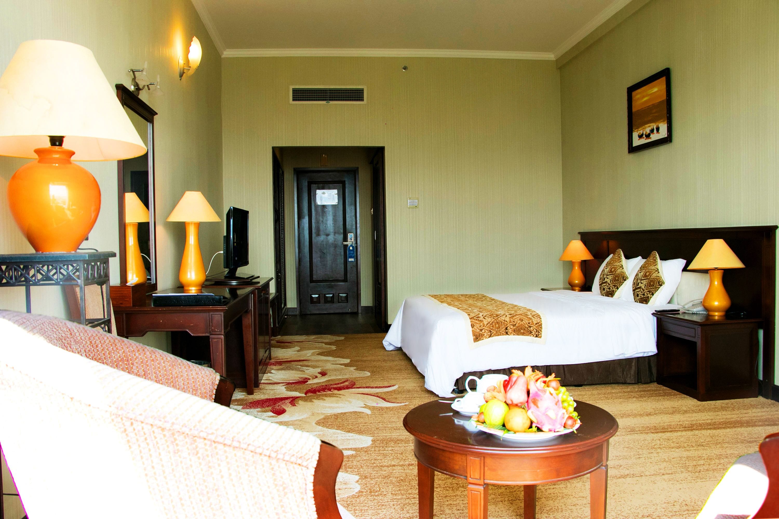 A hotel room with a bed, two bedside tables with lamps, a desk with a lamp, a chair, a small table with fruit, and a door leading to the hallway.