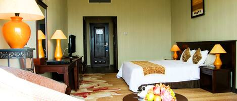 A hotel room with a bed, two bedside tables with lamps, a desk with a lamp, a chair, a small table with fruit, and a door leading to the hallway.