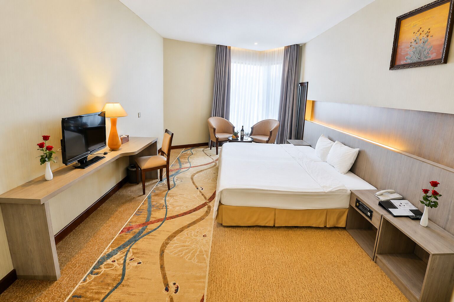 A hotel room with a bed, a desk with a television, a chair, a small table with a bottle, and a painting on the wall.