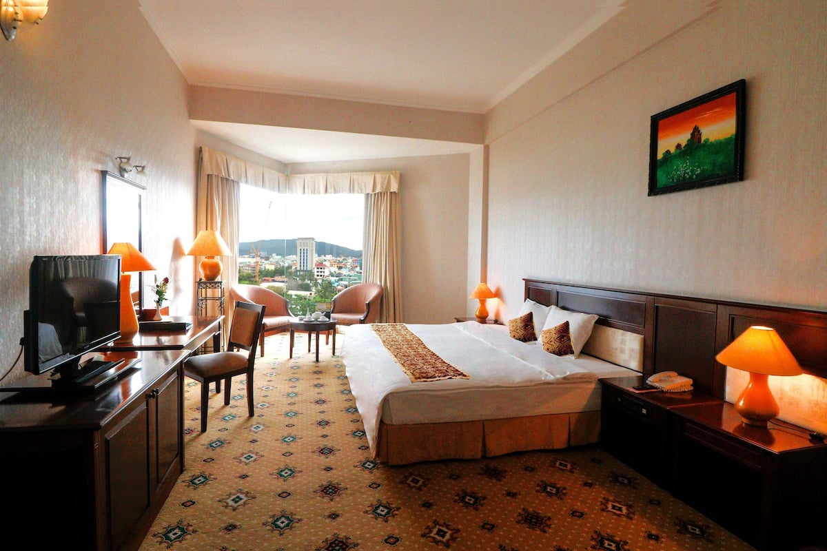 Une chambre d’hôtel avec un lit, un bureau, une télévision, des chaises, des lampes et un tableau au mur.