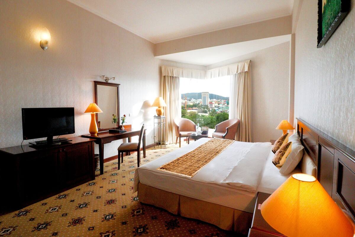 Une chambre d’hôtel équipée d’un lit, d’un bureau, d’une télévision, de deux lampes et offrant une vue sur le paysage urbain par la fenêtre.