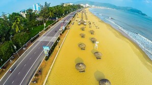 Una playa con filas de tumbonas y sombrillas, una carretera paralela a la playa, y edificios al fondo.