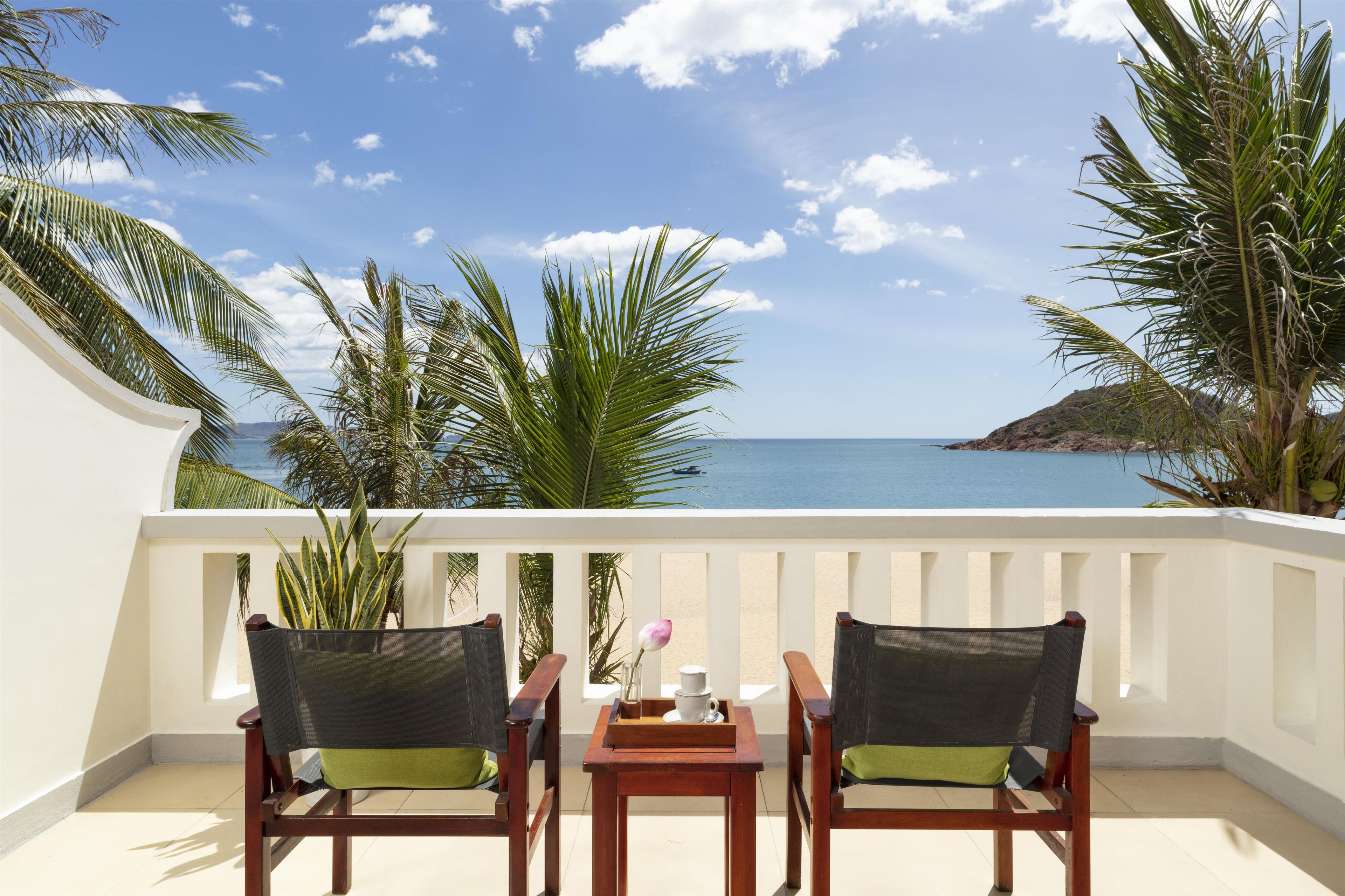 A balcony with two chairs and a table, overlooking a beach with palm trees and the sea.