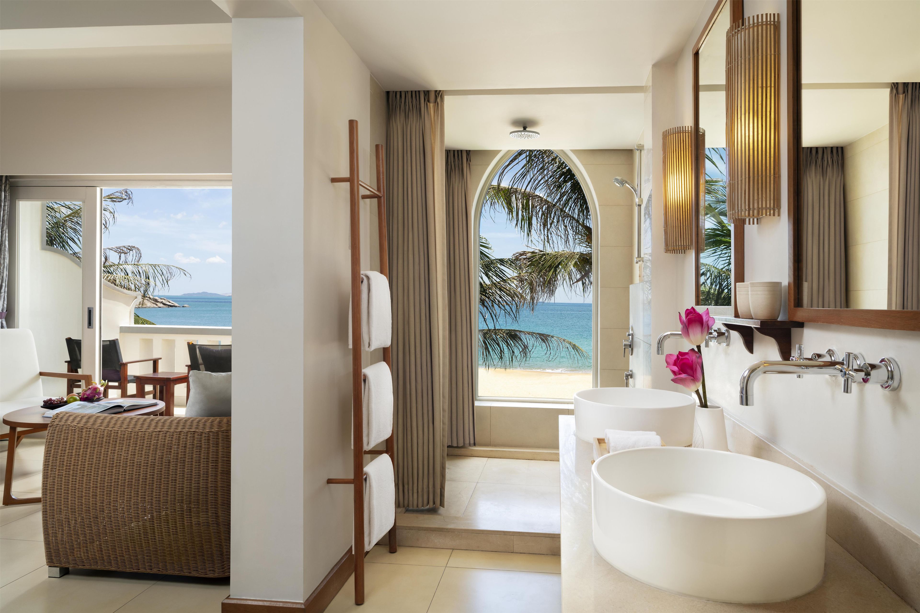 A modern bathroom with a large bathtub, a round sink, and a mirror. There is a view of the beach through a window.