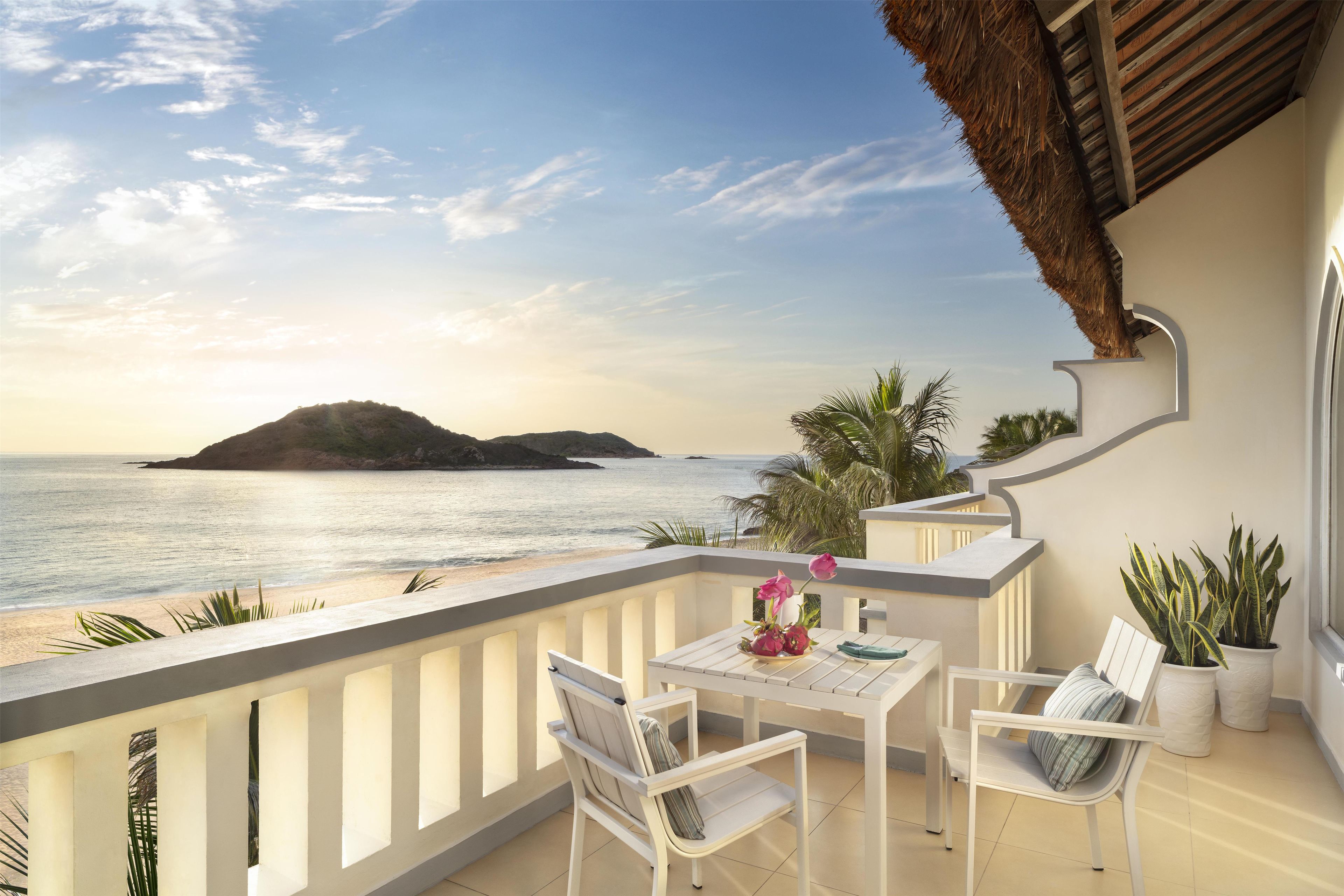 Un balcon avec vue sur la mer et une île lointaine, aménagé avec une table et des chaises, ainsi que des plantes en pot.