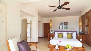 A hotel room with a bed, a chair, a table with a tray of fruit, a ceiling fan, and a brick wall with framed pictures. - Avani Quy Nhon Resort (Quy Nhon Nam)