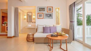 A modern living room with a sofa, a coffee table, and a dining table. There are framed pictures on the wall, a mirror, and a view of the outdoors through large windows. - Avani Quy Nhon Resort (Quy Nhon Nam)