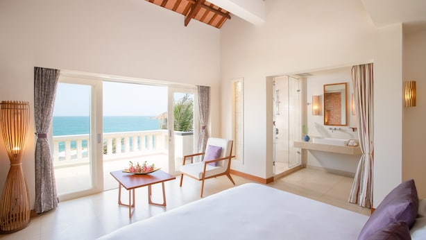 A hotel room with a bed, a chair, a table, a bathroom with a sink, and a view of the sea. - Avani Quy Nhon Resort (Quy Nhon Nam)