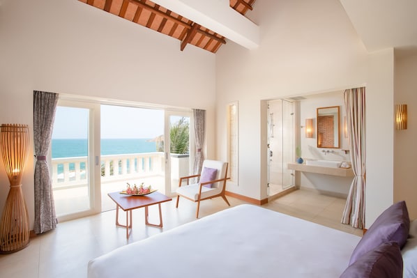 Habitación de hotel con cama, una silla, una mesa, baño con lavabo y vista al mar.