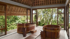 A spa area with wooden tubs and a thatched roof, surrounded by large windows offering a view of lush greenery. - Avani Quy Nhon Resort (Quy Nhon Nam)