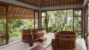 Un área de spa con tinas de madera y techo de paja, rodeada de amplios ventanales con vistas a exuberante vegetación.