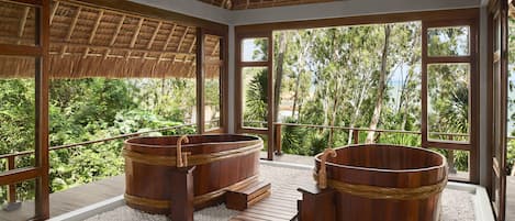 Un área de spa con tinas de madera y techo de paja, rodeada de amplios ventanales con vistas a exuberante vegetación.