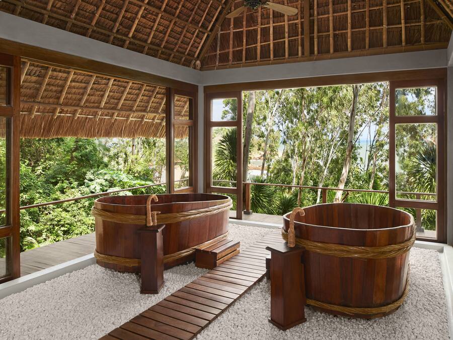 A spa area with wooden tubs and a thatched roof, surrounded by large windows offering a view of lush greenery.
