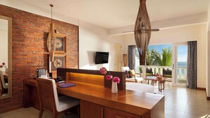 A modern hotel room with a wooden desk, a large window with a view of the ocean, a balcony with a view of palm trees, and a brick accent wall. - Avani Quy Nhon Resort (Quy Nhon Nam)