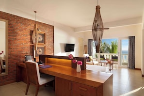 Una habitación de hotel moderna con un escritorio de madera, un ventanal con vista al océano, un balcón con vista a palmeras y una pared de acento de ladrillo.