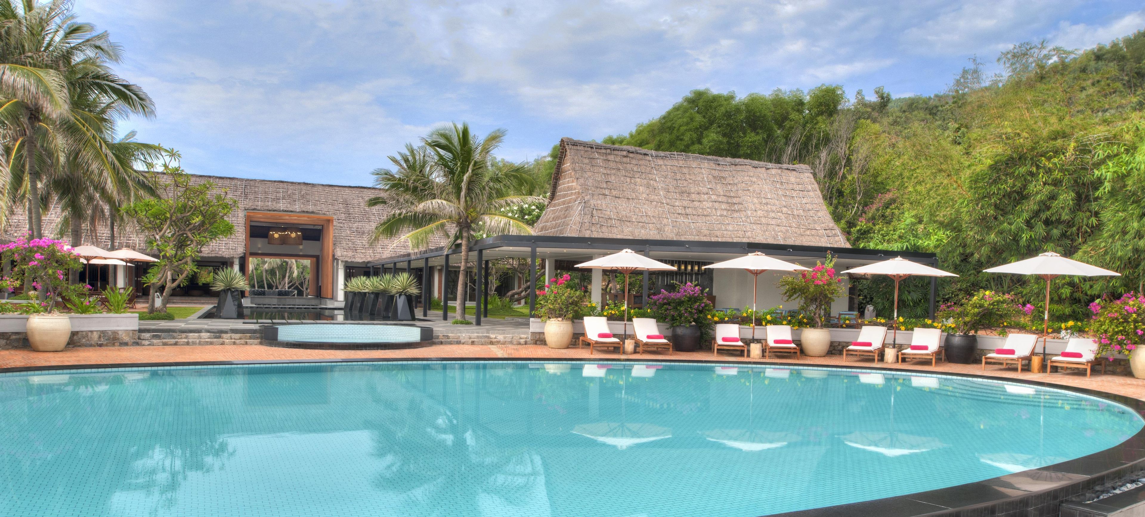 A swimming pool with lounge chairs and umbrellas, surrounded by tropical plants and a building with a thatched roof.