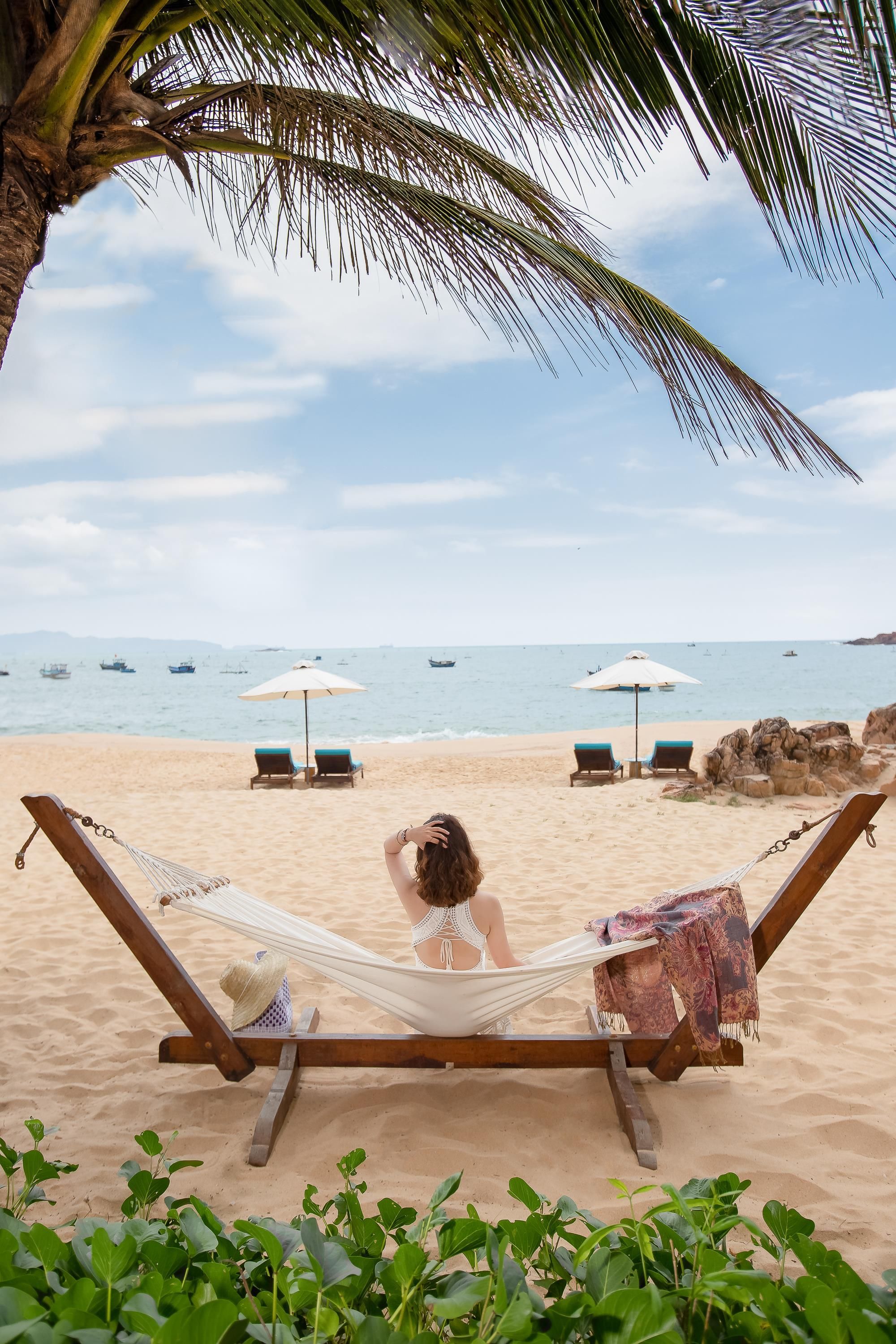 Escena de playa con una hamaca, sillas de descanso y sombrillas bajo una palmera.