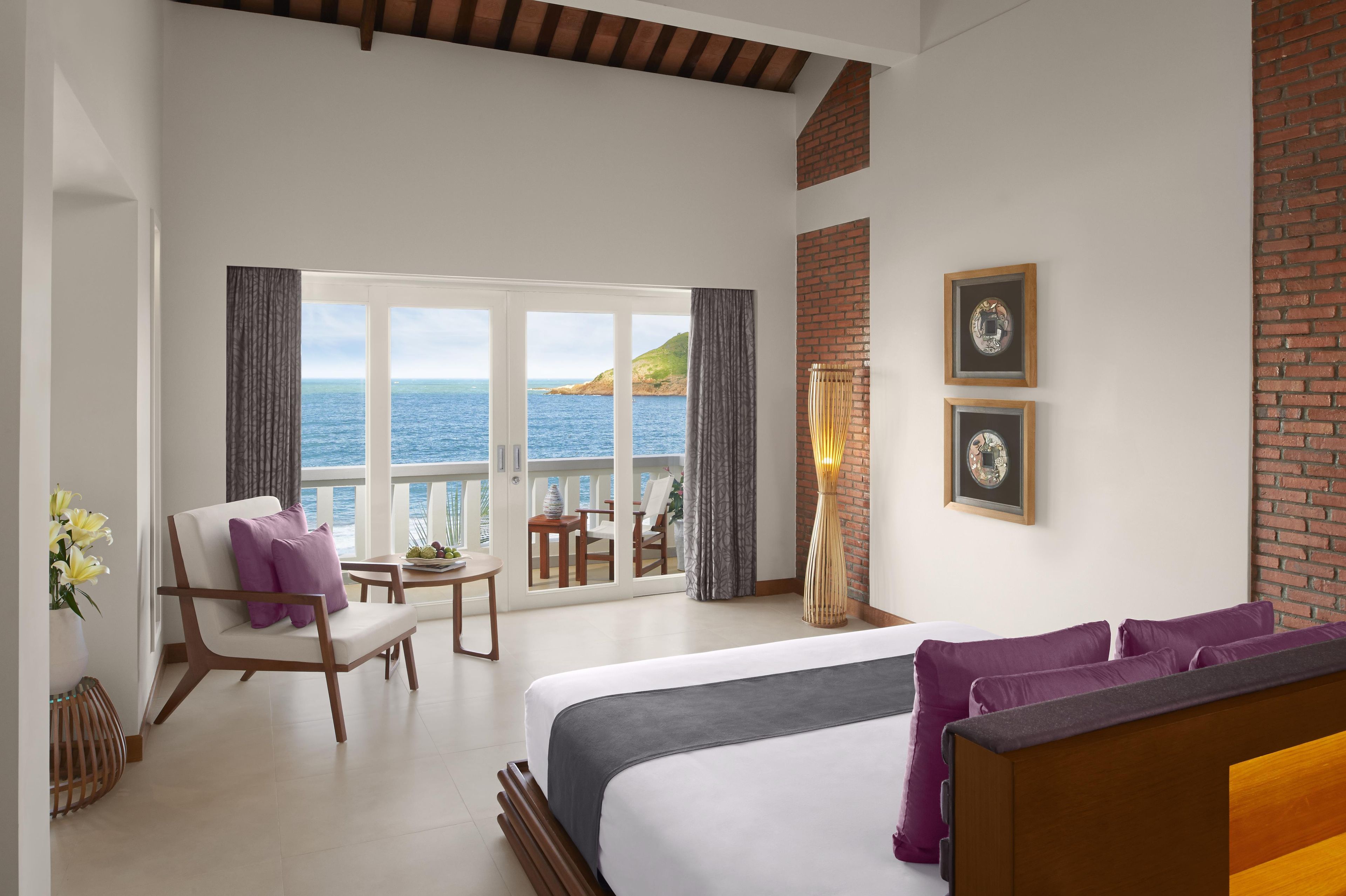 A hotel room with a large bed, a seating area with a table, and a view of the sea through large windows.