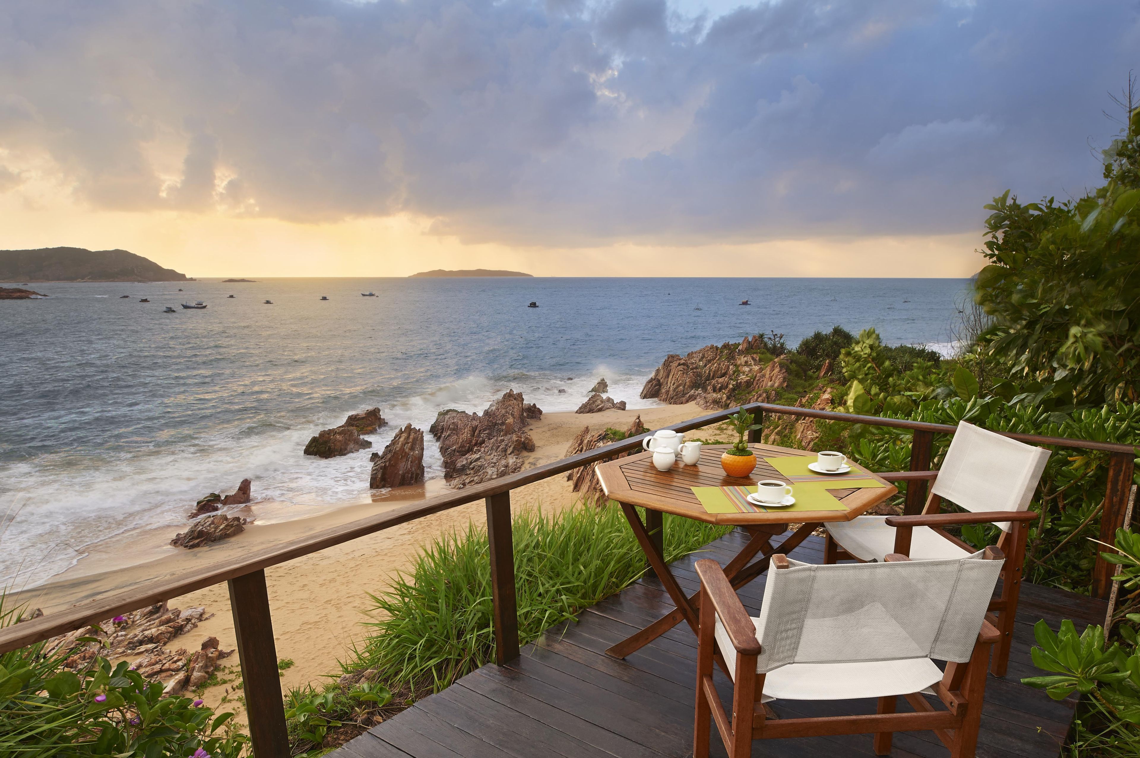 Una terraza de madera con una mesa y sillas con vista a una playa con afloramientos rocosos y el mar.