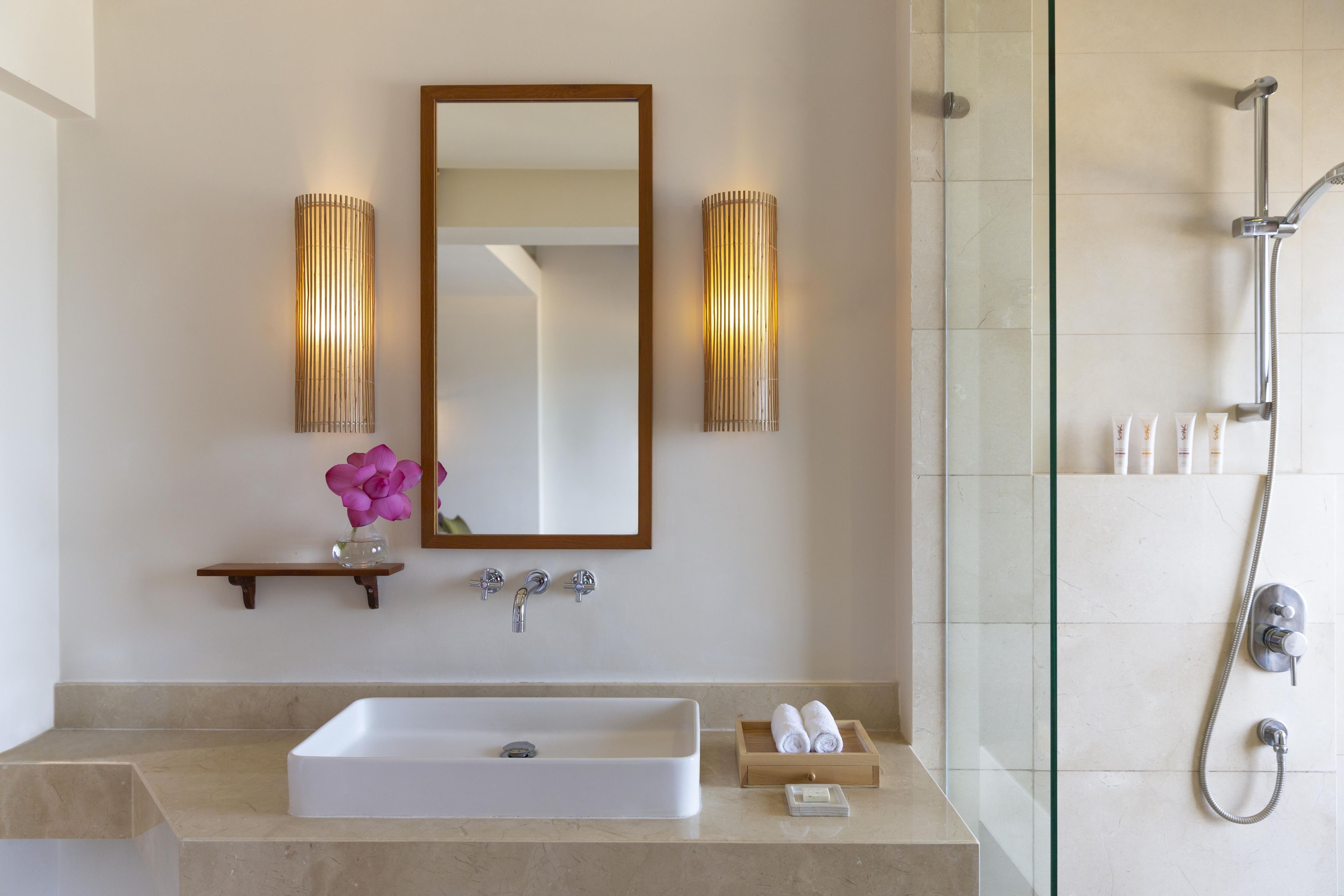 Un bagno con un lavandino rettangolare, uno specchio, due luci a parete, una doccia con soffione manuale e un piccolo vassoio di legno con articoli da toilette.