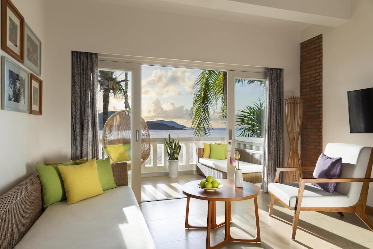 A modern living room with a sofa, armchair, and a table with fruit. A balcony with a view of the sea and mountains is visible.