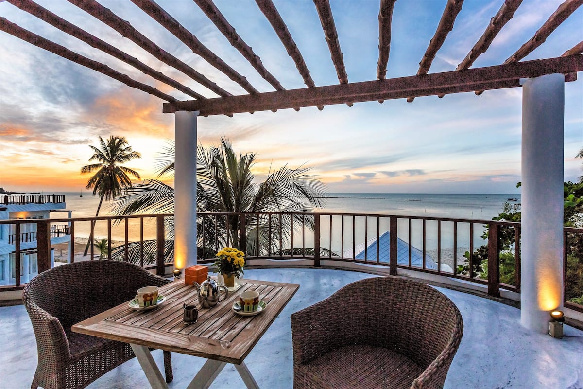 A wooden table with chairs and a view of the ocean at sunset.