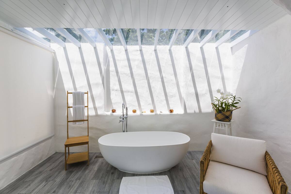 A modern bathroom with a freestanding bathtub, a wooden shelf, a wicker chair, and a potted plant.