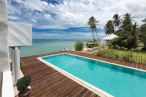 Piscina rectangular con muelle de madera, rodeada de follaje y palmeras, con vista a un mar tranquilo.