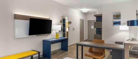A hotel room with a bed, a desk, a chair, a bench, a TV, a mirror, and a lamp.